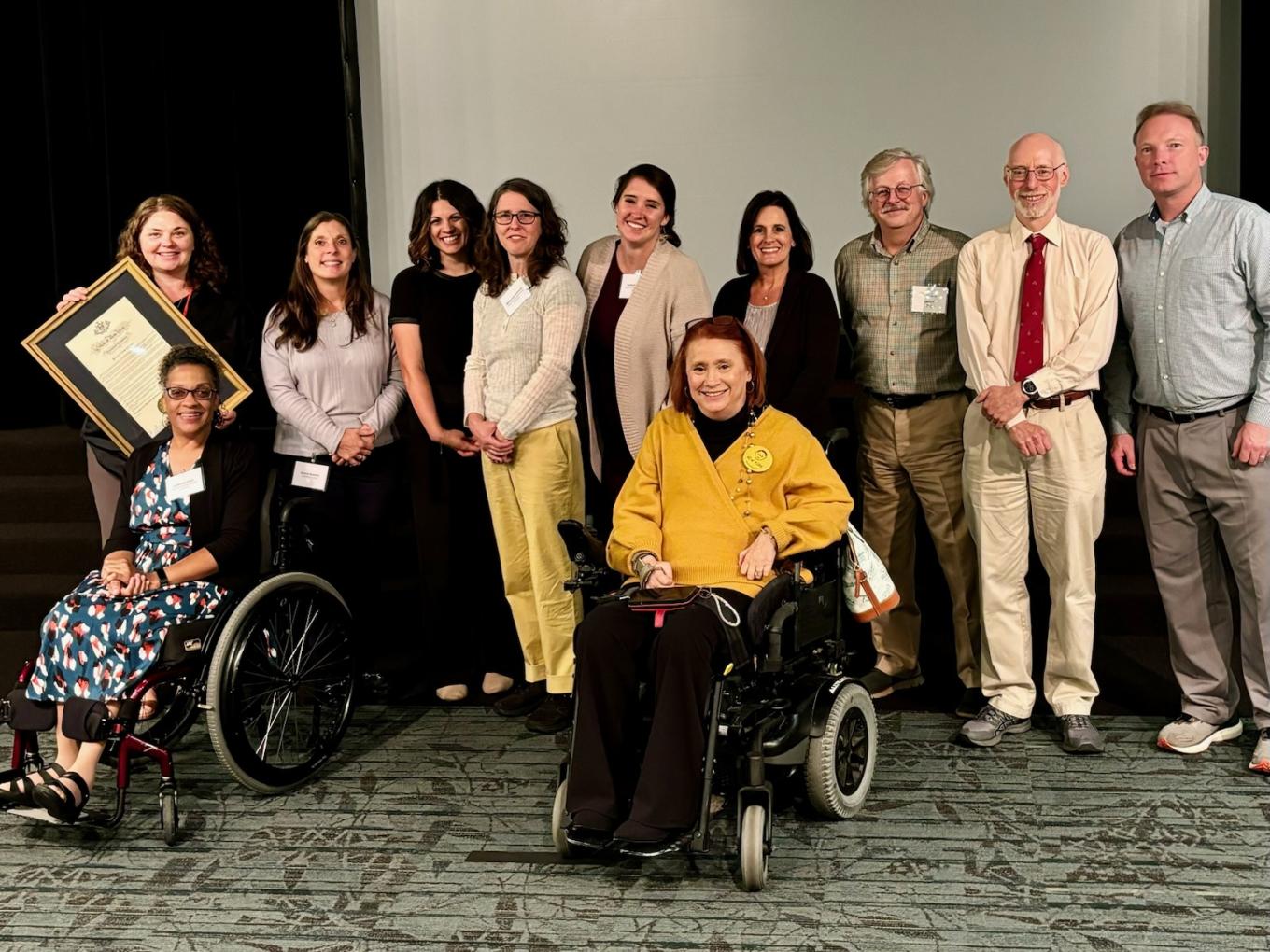 Nine people from the Yang-Tan Institute stand behind two people seated in wheelchairs: one of these seated people is an institute faculty member and the other is Kimberly Hill, New York state’s chief disability officer. Kimberly stands out in the photo because she is wearing a bright yellow sweater. Everyone is smiling and dressed professionally. One person, Ellice Switzer, holds a framed DREAM proclamation; the framed document is almost half as tall as Ellice.