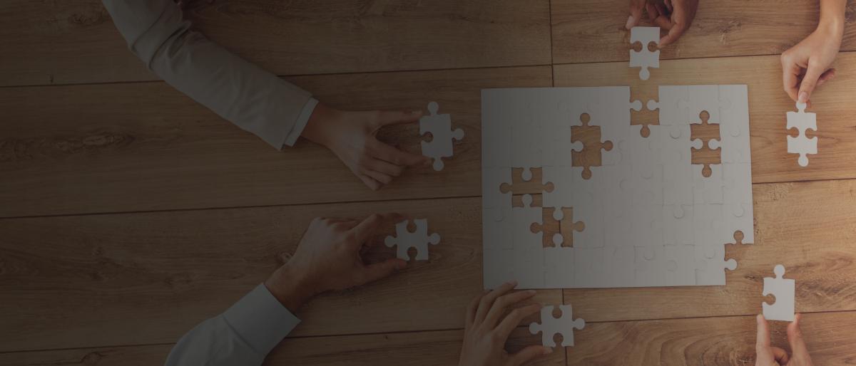 Overhead shot of a puzzle on a wooden table where lots of hands around it hold pieces to the puzzle