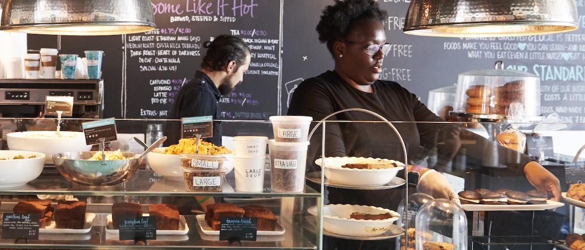 A small business bakery, with staff standing behind a counter. There is a tray of gooey-chocolate-glazed doughnuts.