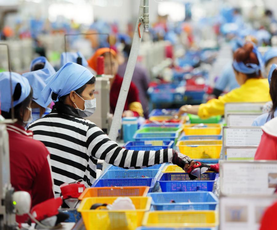 Cambodian workers are seen in a local footwear manufacturing plant, with containers of materials in multiple colors sitting in front of workers wearing bandanas and many wearing masks as well.