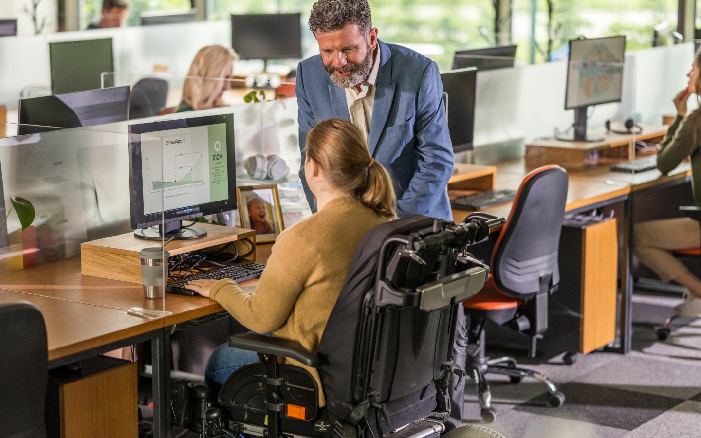 A businessman stands working with a co-worker in a wheelchair, engaging over a computer in a modern office setting