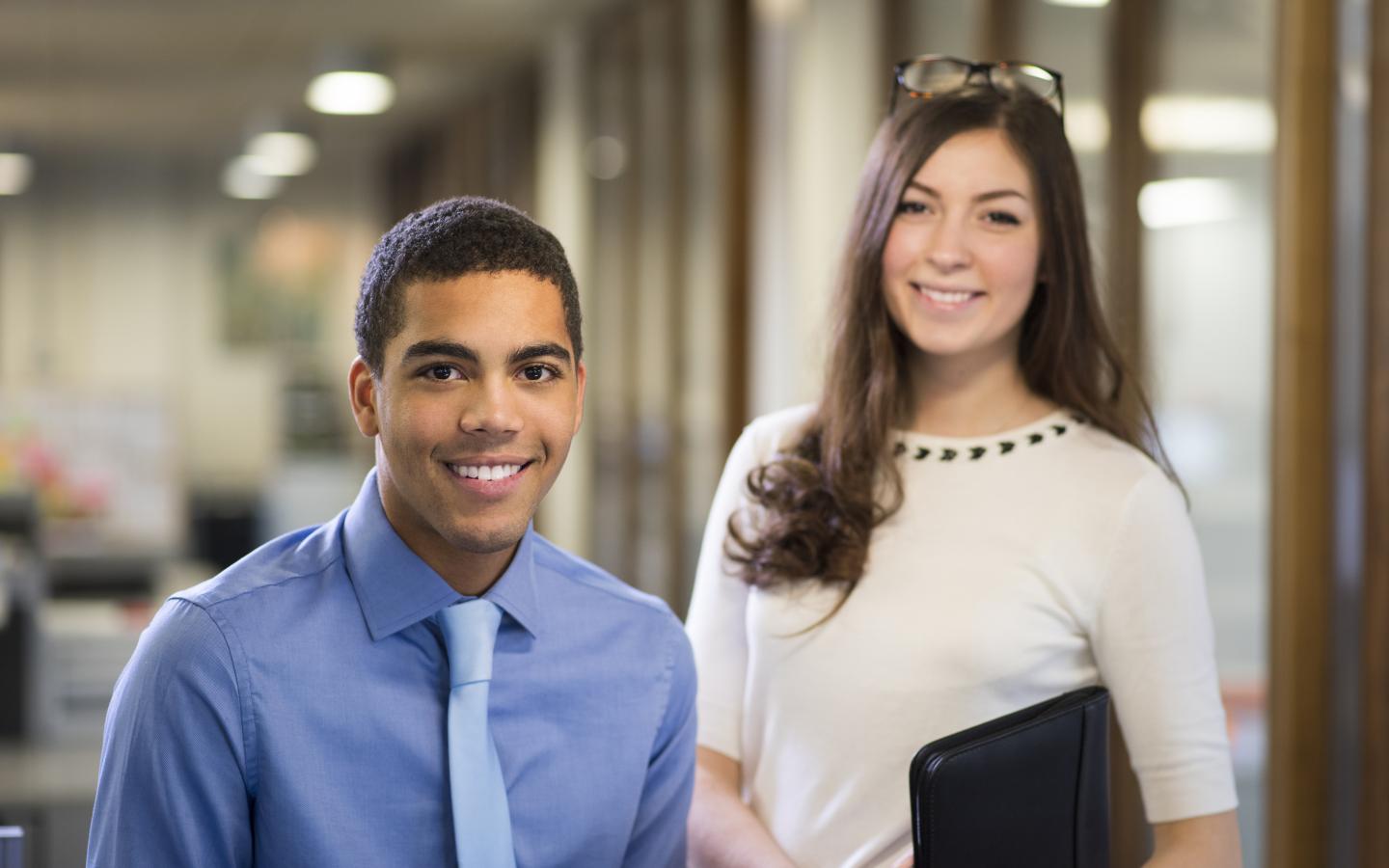 two young people stand in their new workplace smiling proudly to camera