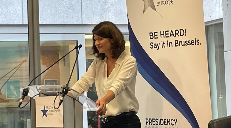 MEP Lara Wolters stands at a lecturn with a Press Club Bussels Europe sign on it, in front of a white banner with black and blue text that says "Press Club Brussels Europe" and "Be Hear. Say it in Brussels" at an event