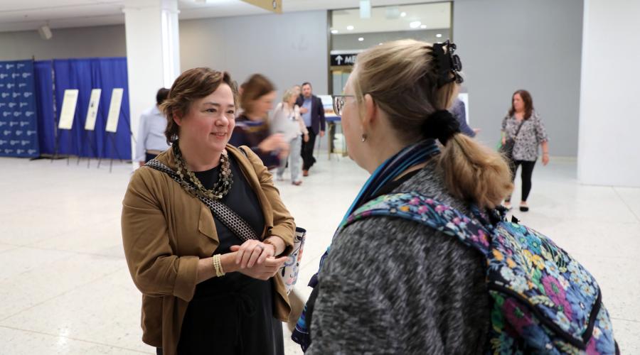 Yang-Tan Institute executive director Wendy Strobel Gower smiles while chatting with a DREAM attendee. Strobel Gower is professionally dressed in chunky jewelry, black dress and tan wrap; the attendee is wearing a blue scarf over a gray top and a backpack with a floral design.