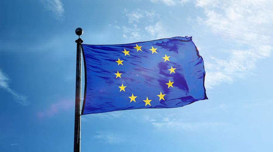 The European Union flag flies in front of a blue sky. The flag is a deep blue, with gold stars representing each of the member nations of the European Union. It is attached to a flag pole.