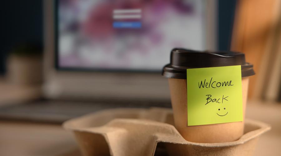 A post-it note that says "Welcome Back" sticks to a take out coffee cup in front of a computer.