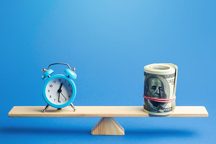 A clock and a roll of hundred dollar bills sit at opposite ends of a scale.