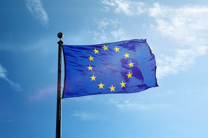 The European Union flag flies in front of a blue sky. The flag is a deep blue, with gold stars representing each of the member nations of the European Union. It is attached to a flag pole.