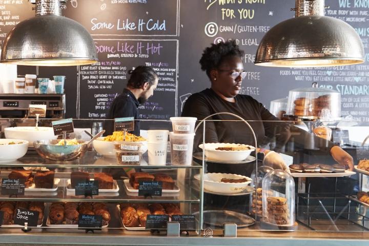 A small business bakery, with staff standing behind a counter. There is a tray of gooey-chocolate-glazed doughnuts.