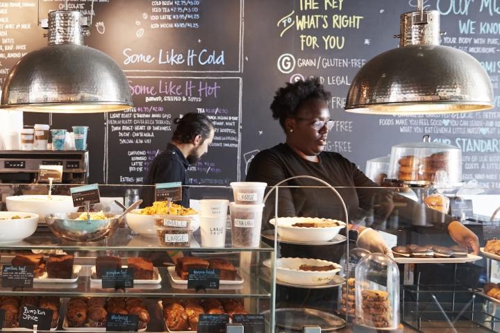 A small business bakery, with staff standing behind a counter. There is a tray of gooey-chocolate-glazed doughnuts.