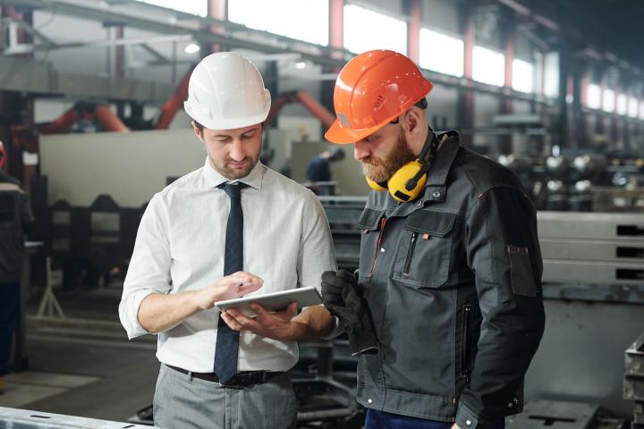 Two workers wearing safety vests and hard hats look over a piece of machinery together. One holds a clipboard, supervising the other.