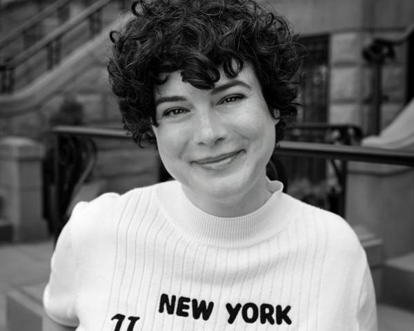 Sarah Krasley, a woman with short curly dark hair, smiles as she wears a shirt that says New York Herald Tribune in a black and white photo