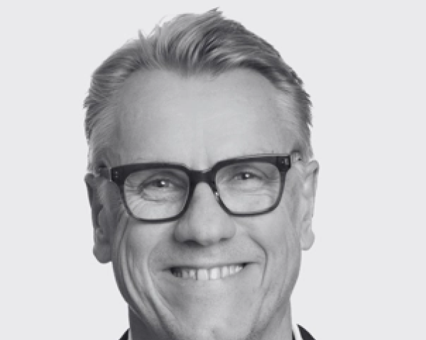 A black and white photo of a Colin Browne smiling as he wears glasses and has his short hair swooped over to his left, wearing a dark blazer and a light colored button-up shirt
