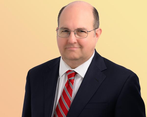 Stephen Vaugn stands wearing a dark suit with a white shirt and a red striped tie. He wears glasses and has hair on the side of his head.