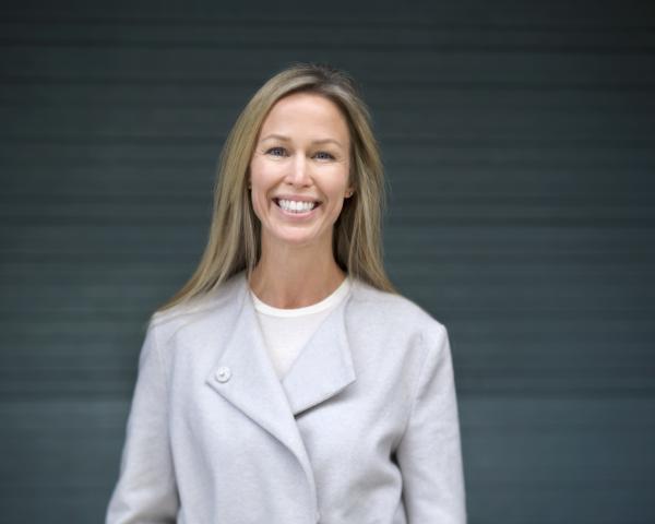 Trude Ertresvåg smiles as she wears a light gray jacket over a white shirt. She has blonde hair, and is standing in front of a gray background.