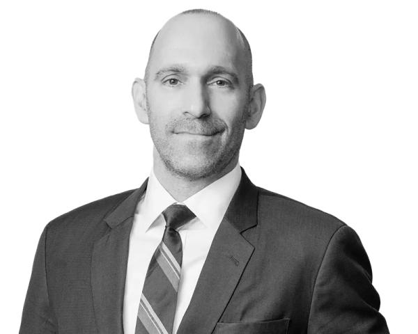Josh Kagan, a man with a bald head, stands and wears a dark suit with a striped tie over a white dress shirt. 