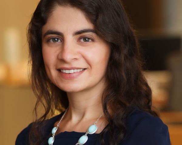 Marina Severinovsky wears a dark dress with a white, peal necklack. She has brown curly hair down to her shoulders.