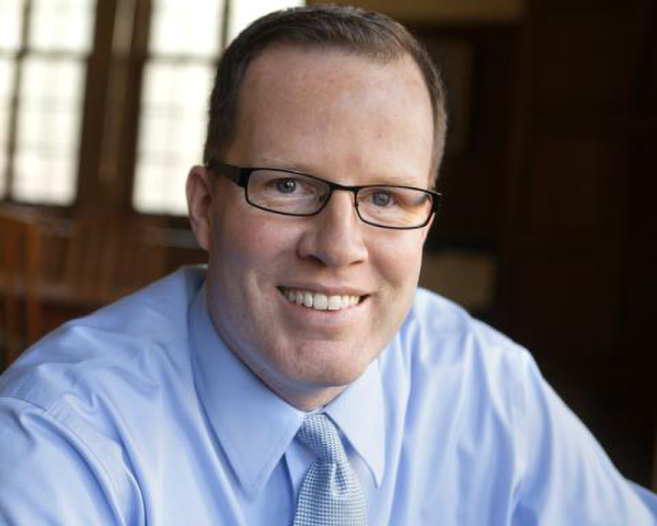 Smiling man wearing glasses, a light blue dress shirt, and a light blue tie, seated indoors near wood-framed windows.
