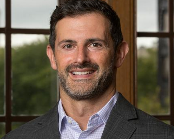 Smiling man with short dark hair and light stubble wearing a gray blazer and light patterned shirt, standing in front of large windows with trees outside.