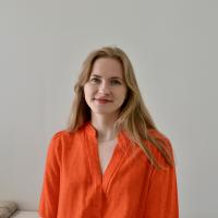 A photo of Victoria Skeie wearing an orange top and standing in front of a white backdrop.