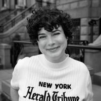 Sarah Krasley, a woman with short curly dark hair, smiles as she wears a shirt that says New York Herald Tribune in a black and white photo