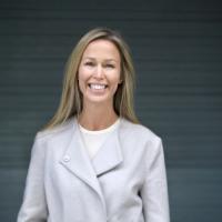 Trude Ertresvåg smiles as she wears a light gray jacket over a white shirt. She has blonde hair, and is standing in front of a gray background.