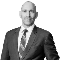 Josh Kagan, a man with a bald head, stands and wears a dark suit with a striped tie over a white dress shirt. 