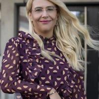 Maria Mexi is a woman with blonde hair and stands outside while wearing a maroon patterned top.