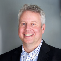 Smiling man with short gray hair wearing a black jacket over a light checkered shirt, posed against a neutral gray studio background.