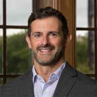 Smiling man with short dark hair and light stubble wearing a gray blazer and light patterned shirt, standing in front of large windows with trees outside.