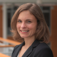 Smiling woman with shoulder-length light brown hair wearing a black blazer, standing indoors near a railing and windows.